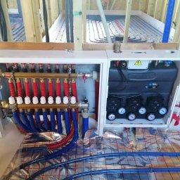 ECO-CIEPEŁKO SPÓŁKA Z OGRANICZONĄ ODPOWIEDZIALNOŚCIĄ - Rozdzielacz ogrzewania podłogowego z czerwono-niebieskimi rurami na folii izolacyjnej, widok z góry, instalacja w trakcie montażu w surowym stanie budynku.