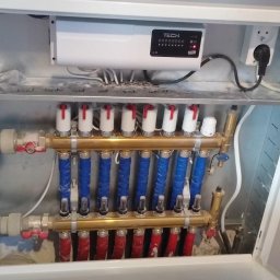 ECO-CIEPEŁKO SPÓŁKA Z OGRANICZONĄ ODPOWIEDZIALNOŚCIĄ - Rozdzielacz ogrzewania podłogowego w szafce, z widocznymi rurami w kolorach niebieskim i czerwonym, oraz sterownikiem TECH z podłączonym zasilaniem.