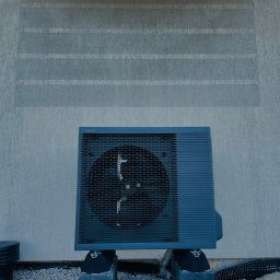 ECO-CIEPEŁKO SPÓŁKA Z OGRANICZONĄ ODPOWIEDZIALNOŚCIĄ - Zamontowana pompa ciepła Panasonic na zewnątrz budynku z szarą elewacją i dwoma oknami. Widoczne elementy instalacji i podłoże z kamyków.