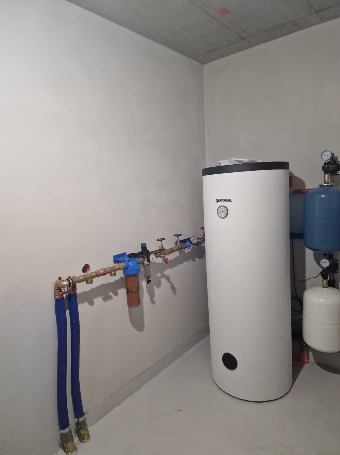 Biały bojler Kospel z podłączonymi rurami i filtrem wody w surowym pomieszczeniu. Widoczne manometry i zawory. Instalacja hydrauliczna w kotłowni.