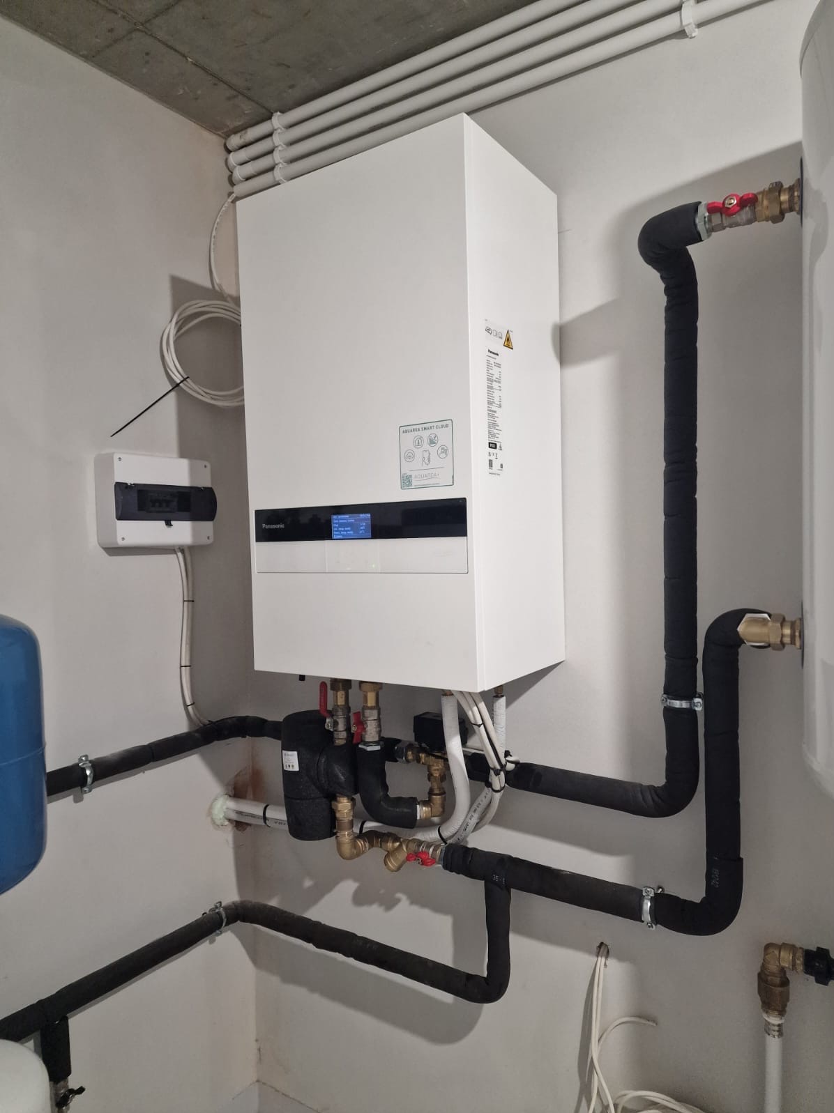 Biała pompa ciepła Panasonic Aquarea Smart Cloud z czarnymi rurami w izolacji, zamontowana na ścianie w pomieszczeniu technicznym. Widoczna skrzynka elektryczna i zbiornik.