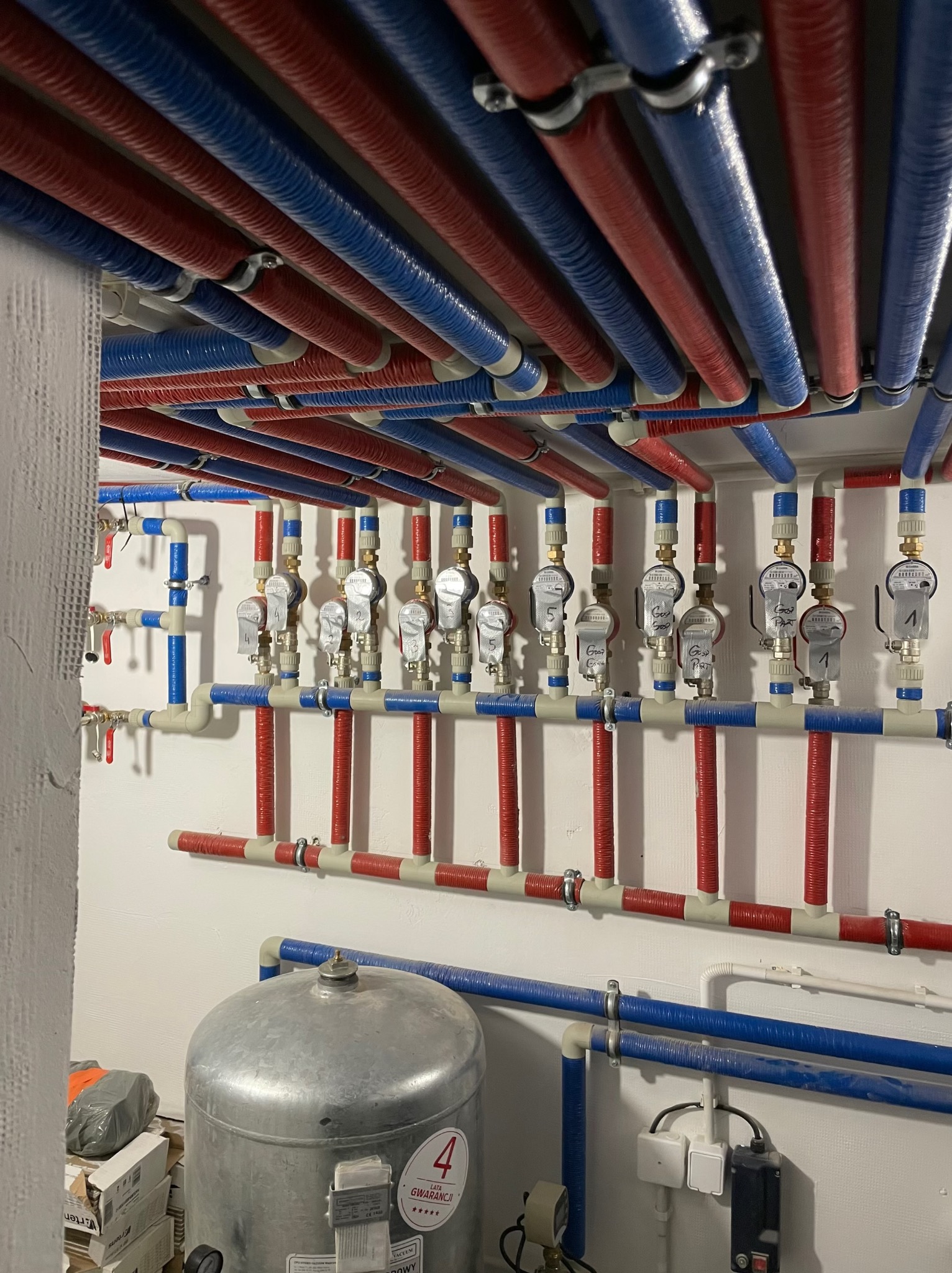 Rozbudowana instalacja grzewcza z rurami w kolorach czerwonym i niebieskim, zaworami i licznikiem. Widoczny zbiornik ciśnieniowy i fragment ściany.
