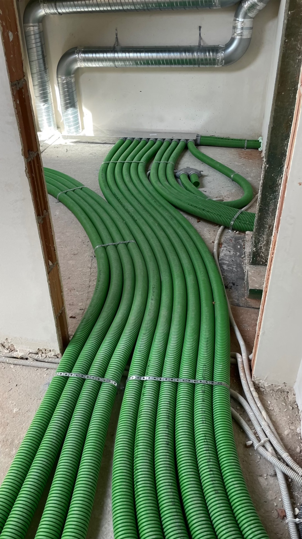 Zielone rury instalacji ogrzewania podłogowego, ułożone równolegle na surowej posadzce, widoczne metalowe mocowania i fragmenty instalacji wentylacyjnej pod sufitem.