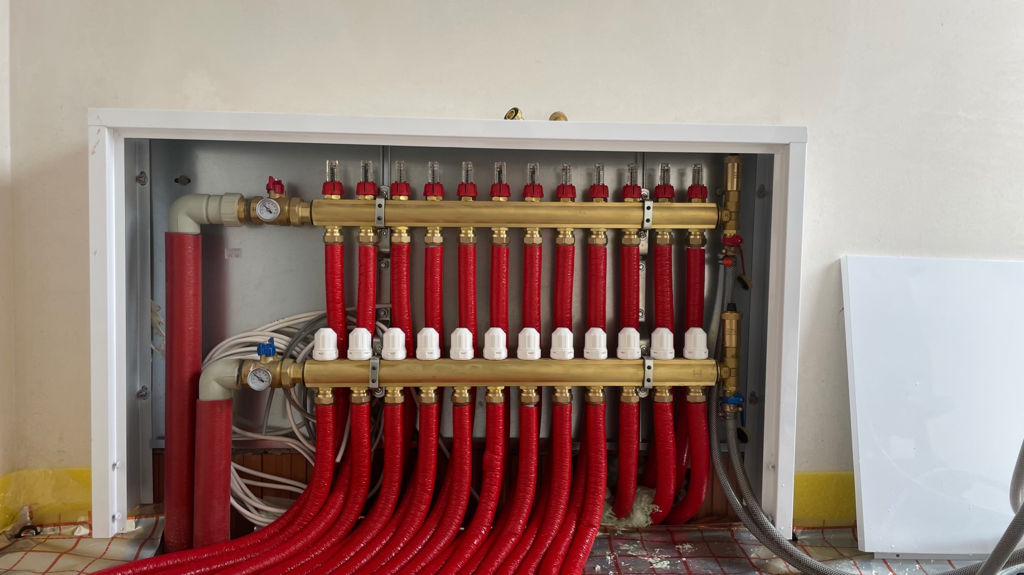 Rozdzielacz ogrzewania podłogowego z czerwonymi rurami w szafce, widoczne manometry i białe regulatory przepływu, ułożony na macie grzewczej.