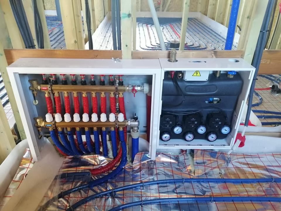 Rozdzielacz ogrzewania podłogowego z czerwono-niebieskimi rurami na folii izolacyjnej, widok z góry, instalacja w trakcie montażu w surowym stanie budynku.