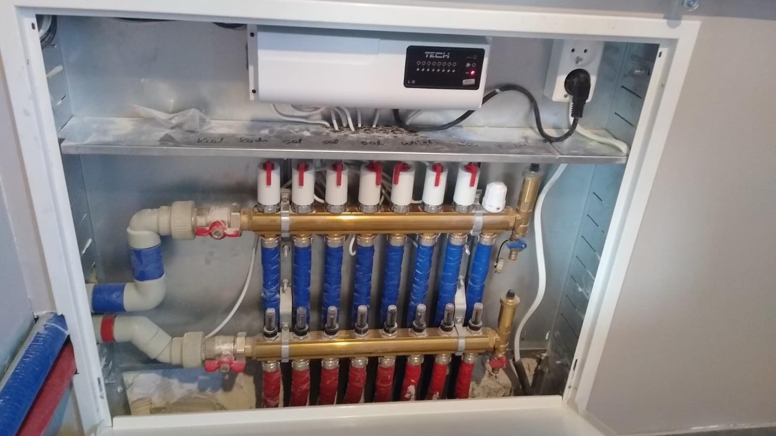 Rozdzielacz ogrzewania podłogowego w szafce, z widocznymi rurami w kolorach niebieskim i czerwonym, oraz sterownikiem TECH z podłączonym zasilaniem.