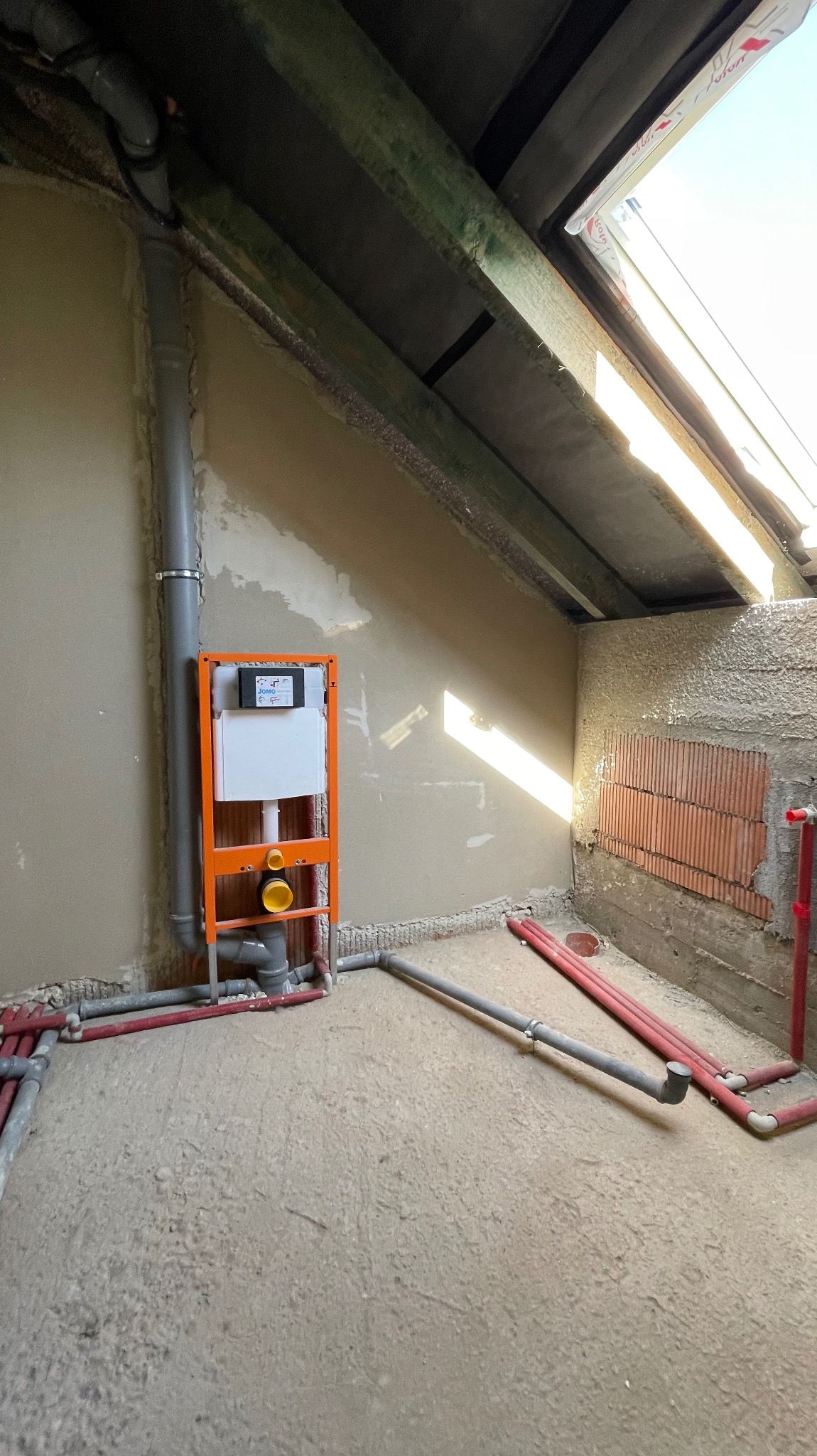 Skośne poddasze z instalacją podtynkową WC w pomarańczowej ramie, szare rury kanalizacyjne i czerwone rury wodne na surowej posadzce. Widoczne cegły i okno dachowe.