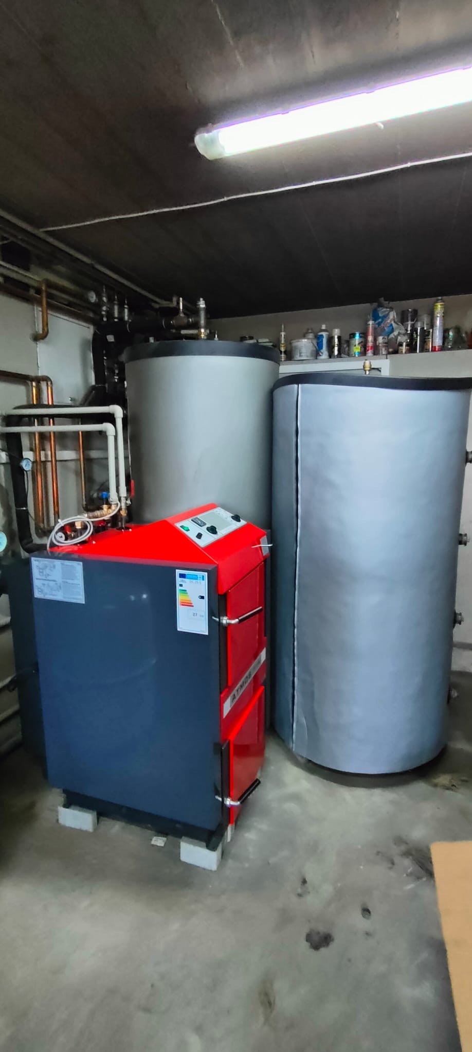 Czerwono-szary piec Atmos na betonowych bloczkach w kotłowni, obok dwa szare zbiorniki akumulacyjne. Widoczne rury instalacji grzewczej i półka z akcesoriami.