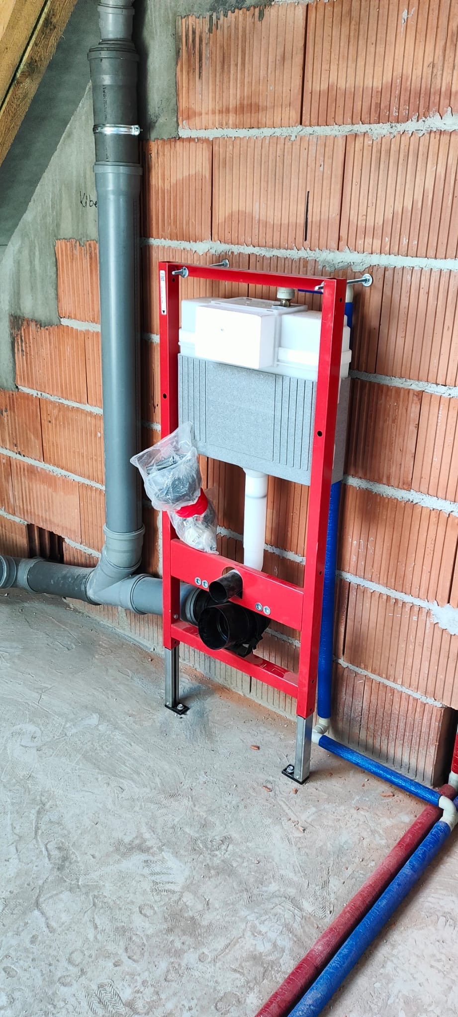 Czerwony stelaż podtynkowy WC z szarą spłuczką, zamontowany przy ceglanej ścianie w surowym stanie. Widoczne szare rury kanalizacyjne i niebiesko-czerwone rury doprowadzające wodę.