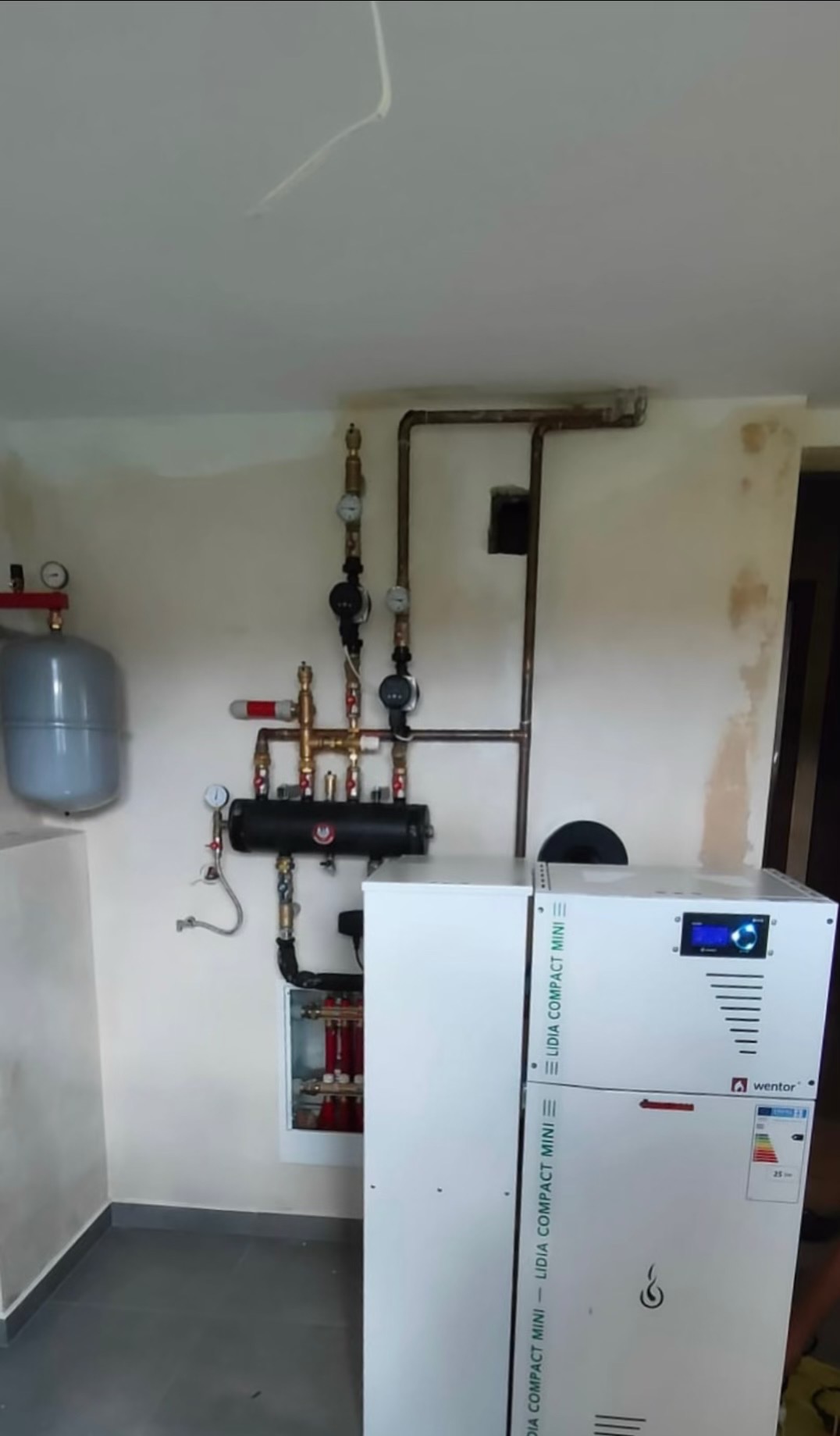Biały piec gazowy Wentor Lidia Compact Mini z instalacją hydrauliczną na ścianie w pomieszczeniu z szarą podłogą i fragmentami nieotynkowanej ściany.