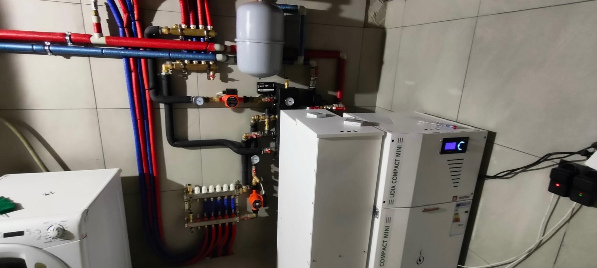 Złożona instalacja grzewcza z piecem gazowym Lidia Compact Mini, rurami w izolacji czerwonej i niebieskiej, rozdzielaczem i pompą obiegową na tle płytek ceramicznych.