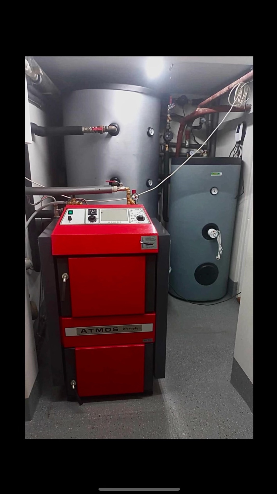 Czerwony piec Atmos Drevoplyn w kotłowni, obok zbiorniki akumulacyjne. Widoczne rury i zawory instalacji grzewczej. Szare ściany i podłoga.