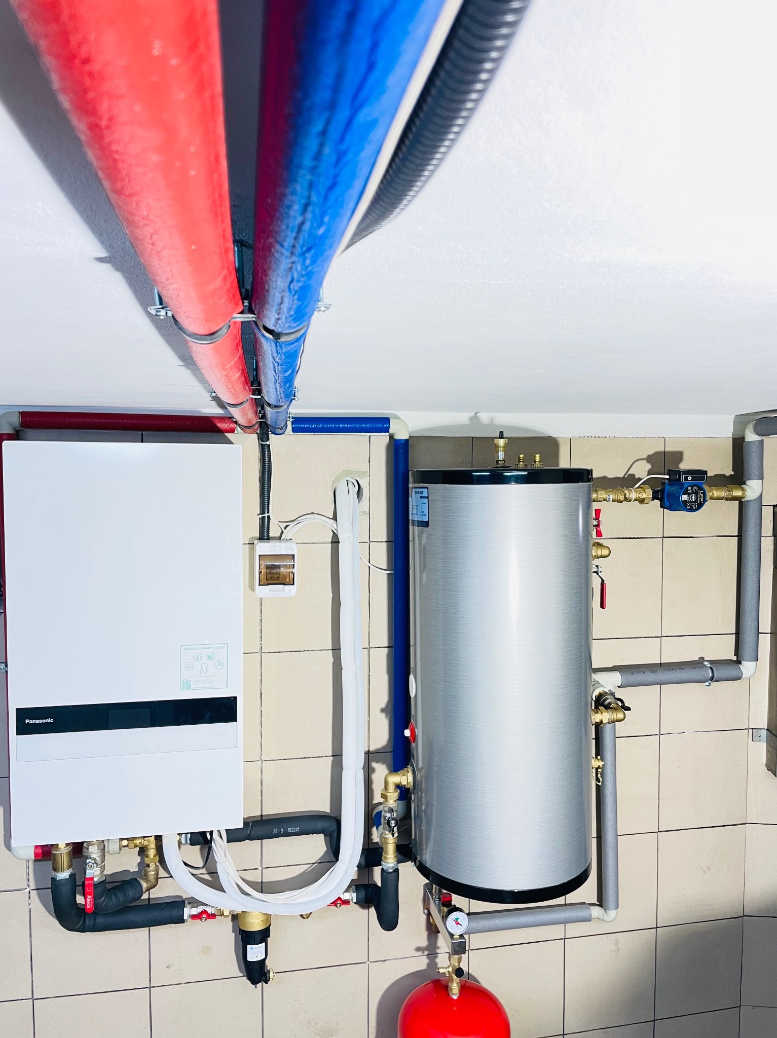 Nowoczesna instalacja grzewcza z piecem gazowym Panasonic i zasobnikiem ciepłej wody, rury w kolorach niebieskim i czerwonym, na tle kafelkowanej ściany.