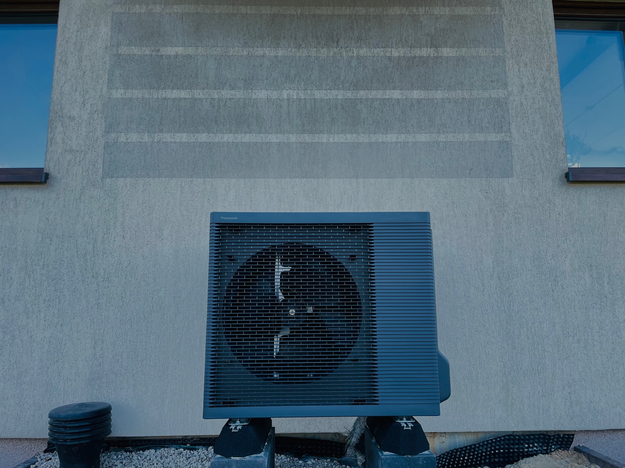 Zamontowana pompa ciepła Panasonic na zewnątrz budynku z szarą elewacją i dwoma oknami. Widoczne elementy instalacji i podłoże z kamyków.
