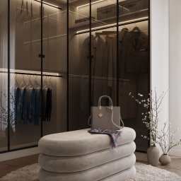 GES STUDIO Wykończenia wnętrz Sylwia i Wiktor Gęs - Nowoczesna garderoba z przesuwanymi szklanymi drzwiami, oświetleniem LED, ubraniami na wieszakach i półkach, z beżową pufą na pierwszym planie, na której leży torebka i apaszka w kwiaty.