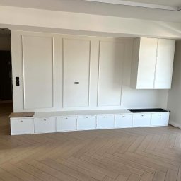 GES STUDIO Wykończenia wnętrz Sylwia i Wiktor Gęs - Biała zabudowa meblowa z szafkami i siedziskiem w salonie, parkiet ułożony w jodełkę, minimalistyczny design, jasne wnętrze z oknem balkonowym.