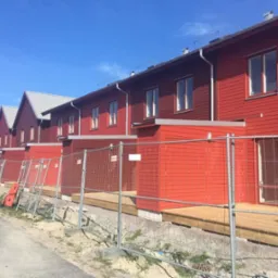 Rząd nowych, czerwonych domów z drewnianą elewacją, zabezpieczonych tymczasowym ogrodzeniem z siatki.