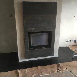 Odynets Aleksandr - Obudowa kominka z ciemnych, matowych płytek, z widocznym wkładem kominkowym. Podłoga zabezpieczona kartonem, ściany w trakcie prac wykończeniowych.