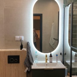 Odynets Aleksandr - Nowoczesna łazienka z owalnym lustrem LED, beżowymi płytkami i drewnianą szafką. Widoczna kabina prysznicowa z ciemnozielonymi płytkami i złotą armaturą.