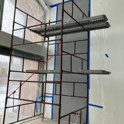 Odynets Aleksandr - Wnętrze w trakcie malowania: rusztowanie na tle ściany zabezpieczonej folią i taśmą malarską. Widoczne okno i przygotowane podłoże. Etap prac wykończeniowych.