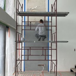 Odynets Aleksandr - Pracownik na rusztowaniu szpachluje ścianę w pomieszczeniu z dużymi oknami, zabezpieczonymi folią. Na podłodze widoczne taśmy malarskie.