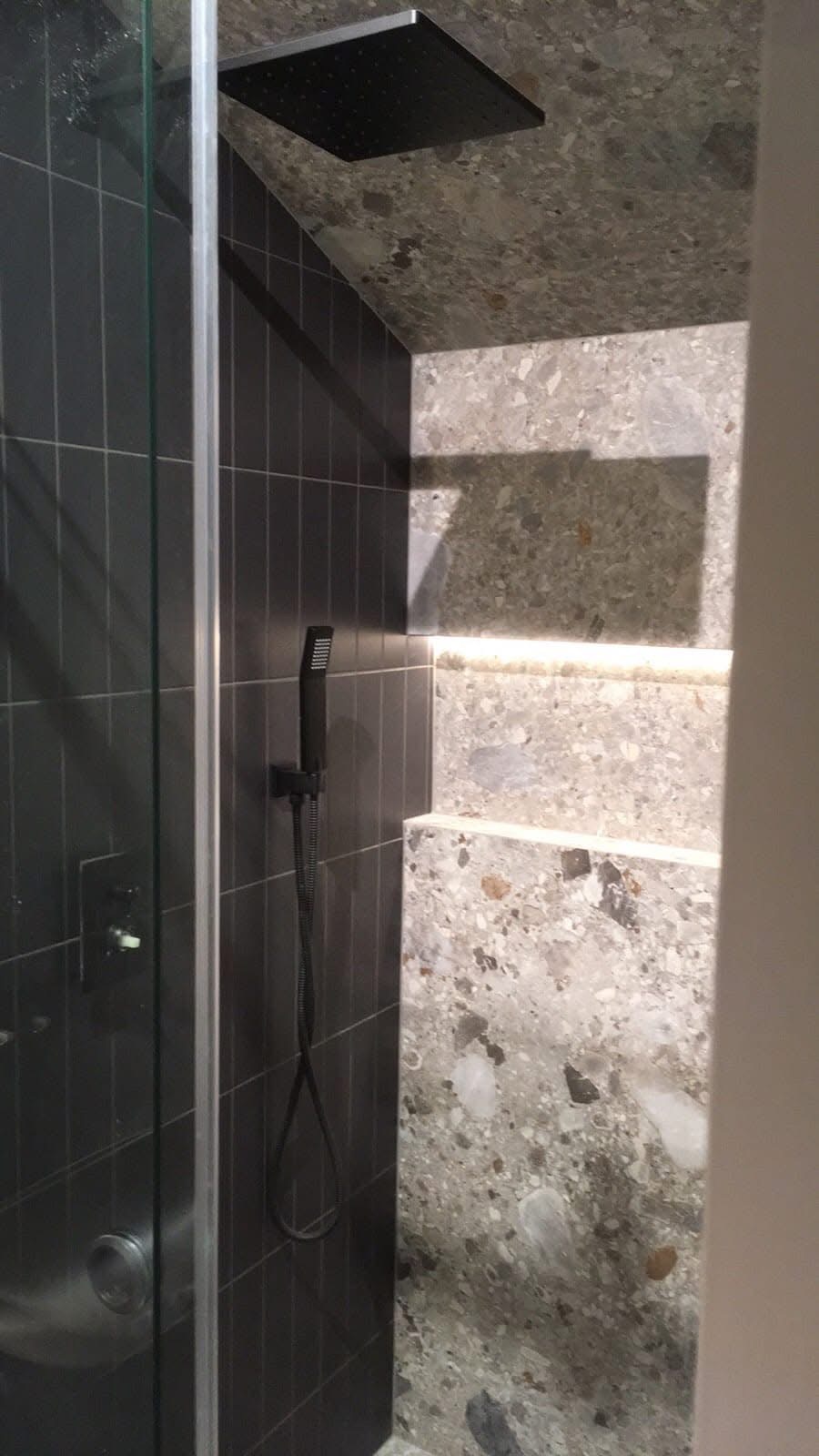 Nowoczesna kabina prysznicowa z czarnymi płytkami i deszczownicą, wykończona mozaiką na ścianie. Widoczny szklany panel i bateria. Minimalistyczny design.