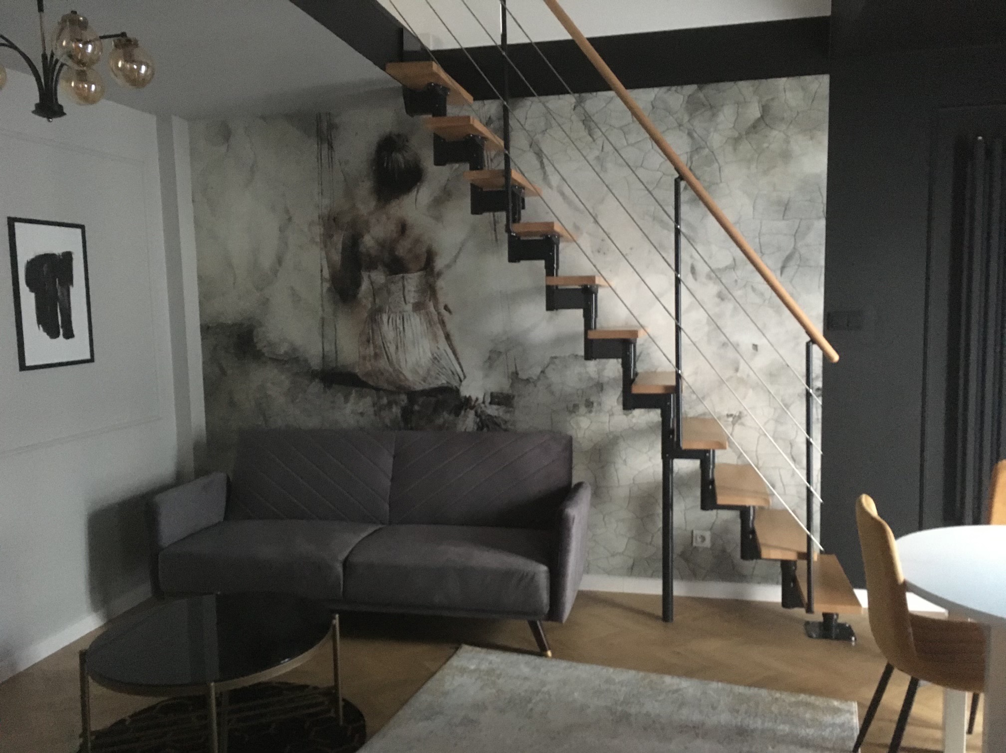 Nowoczesne wnętrze: szara sofa, szklany stolik, schody z drewnianymi stopniami i metalowymi barierkami na tle ściany z artystycznym wzorem. Minimalistyczny design.