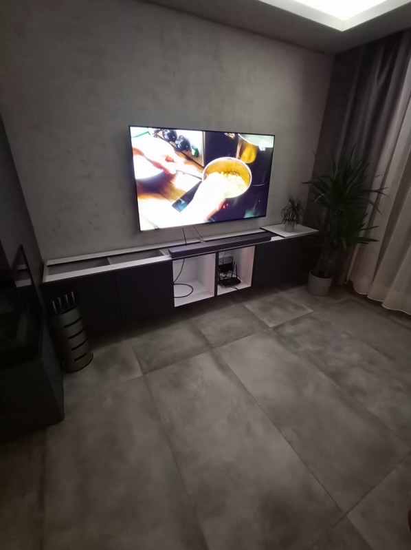 Nowoczesny salon z szarą ścianą, podświetlaną szafką RTV i dużym telewizorem. Minimalistyczny design, płytki podłogowe imitujące beton, roślina doniczkowa w rogu.