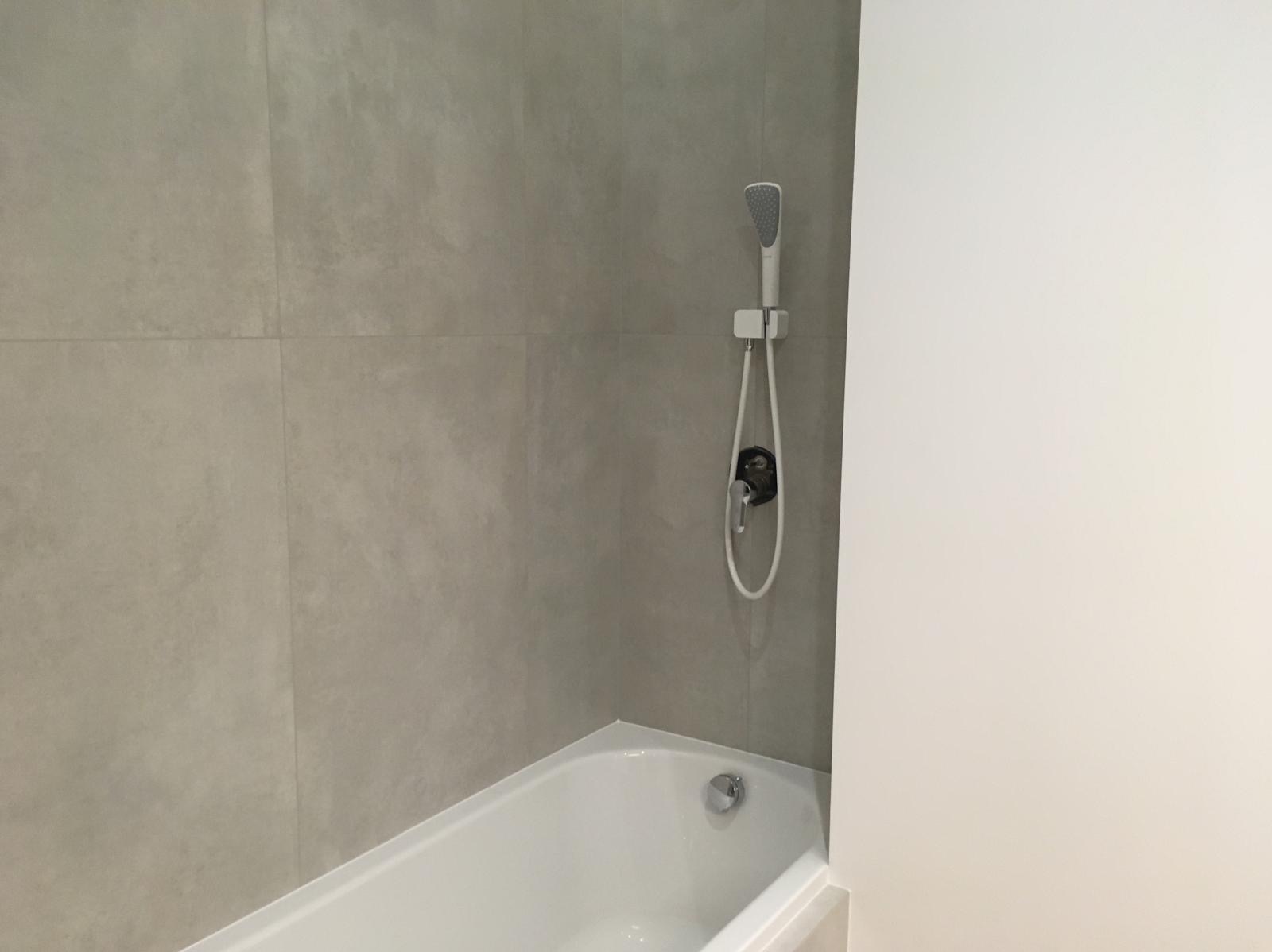 Minimalistyczna łazienka: szare płytki ścienne, biała wanna i chromowana bateria prysznicowa. Nowoczesny design, stonowana kolorystyka, czyste linie.