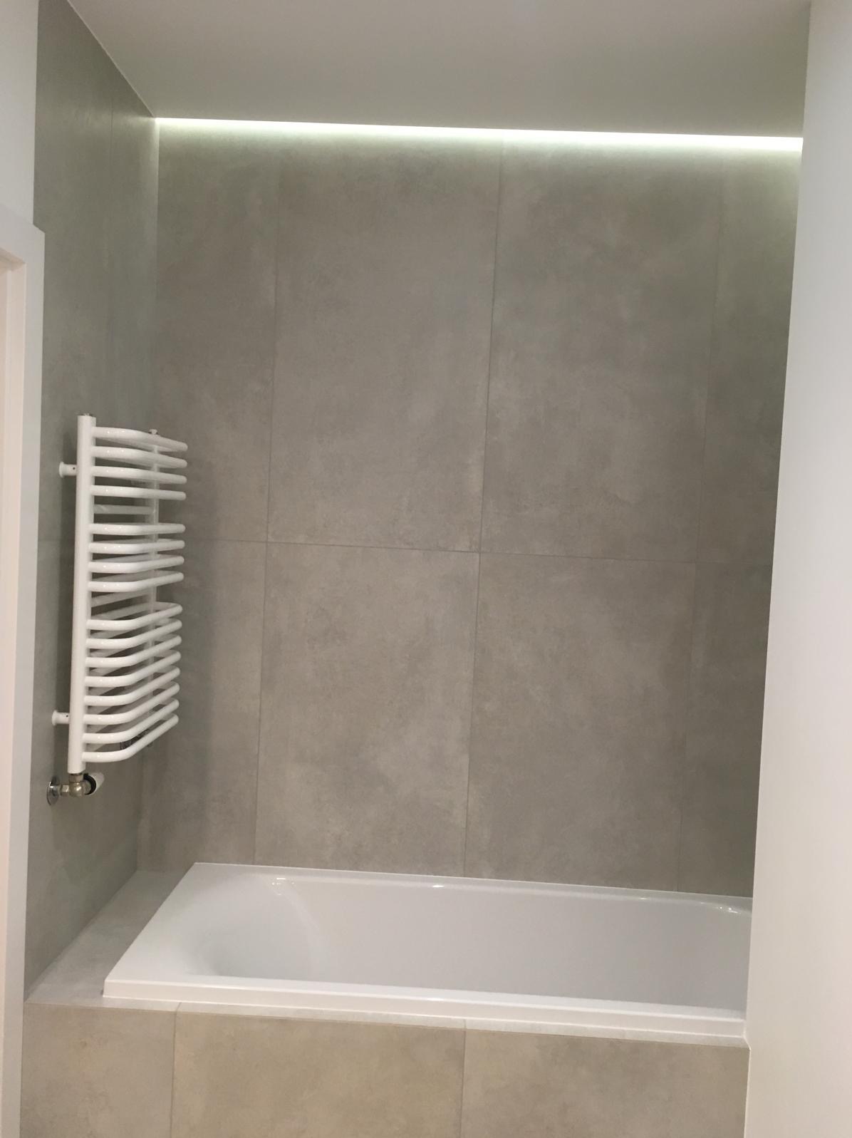 Minimalistyczna łazienka z wanną, grzejnikiem drabinkowym i szarymi płytkami. Podświetlenie LED nadaje nowoczesny charakter. Ujęcie z perspektywy wejścia.