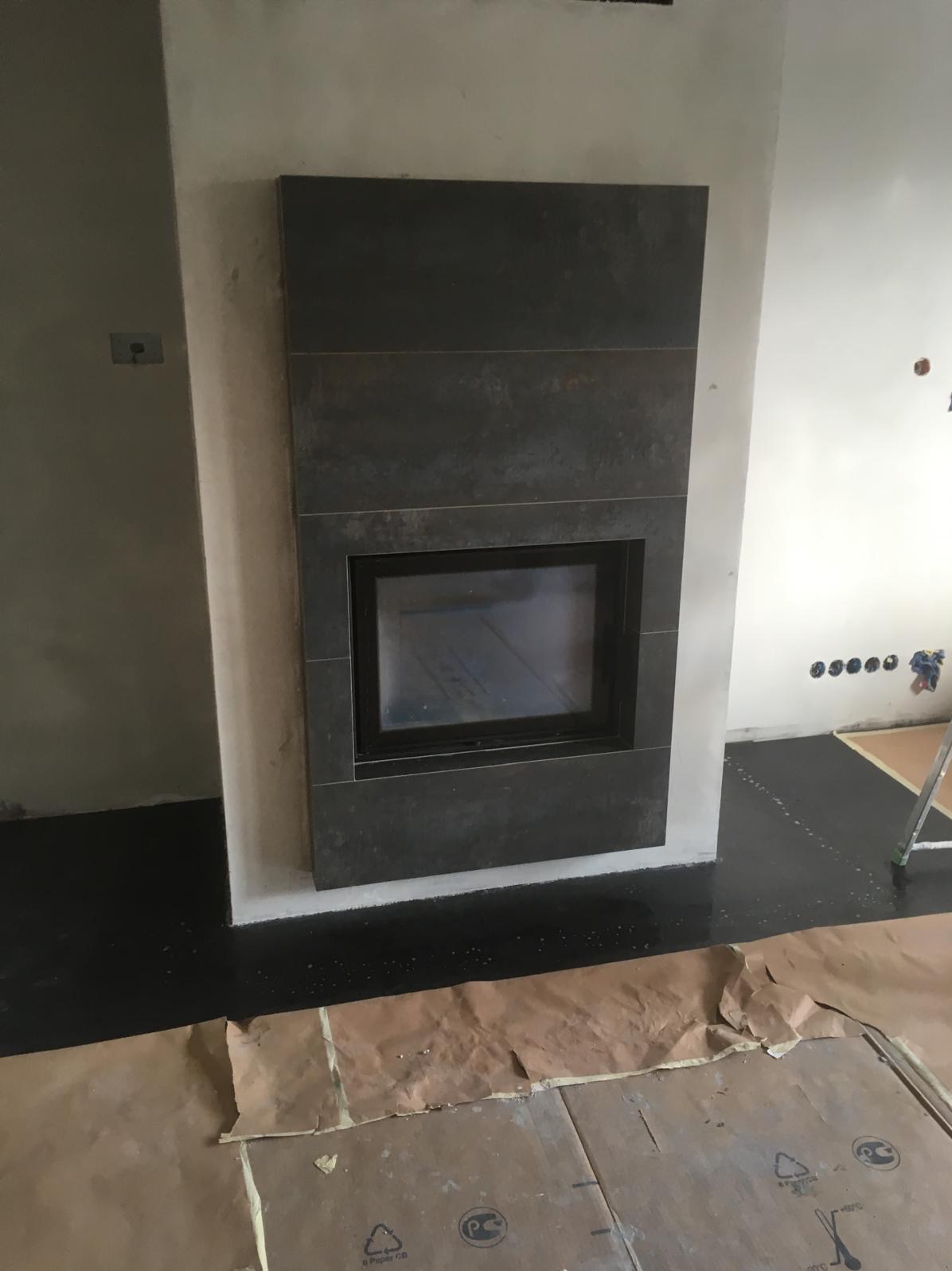 Obudowa kominka z ciemnych, matowych płytek, z widocznym wkładem kominkowym. Podłoga zabezpieczona kartonem, ściany w trakcie prac wykończeniowych.