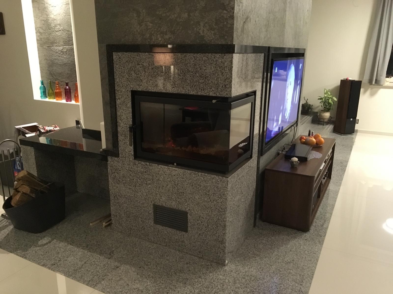 Granitowa obudowa kominka z szybą i wbudowanym telewizorem w salonie. Obok kominka półka z czarnego granitu. W tle dekoracyjne butelki i głośnik.