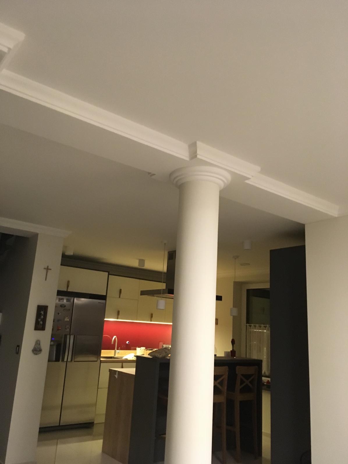 Biała kolumna z dekoracyjnym kapitelem i listwami sufitowymi w jasnym wnętrzu z widoczną kuchnią w tle. Ujęcie z dołu, podkreślające detale architektoniczne.