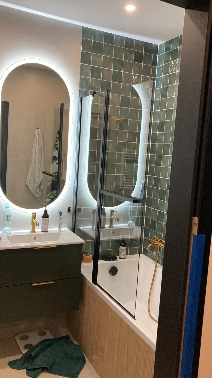 Nowoczesna łazienka z zielonymi płytkami, owalnym lustrem z podświetleniem LED, wanną z parawanem i umywalką w kolorze khaki z złotą armaturą.