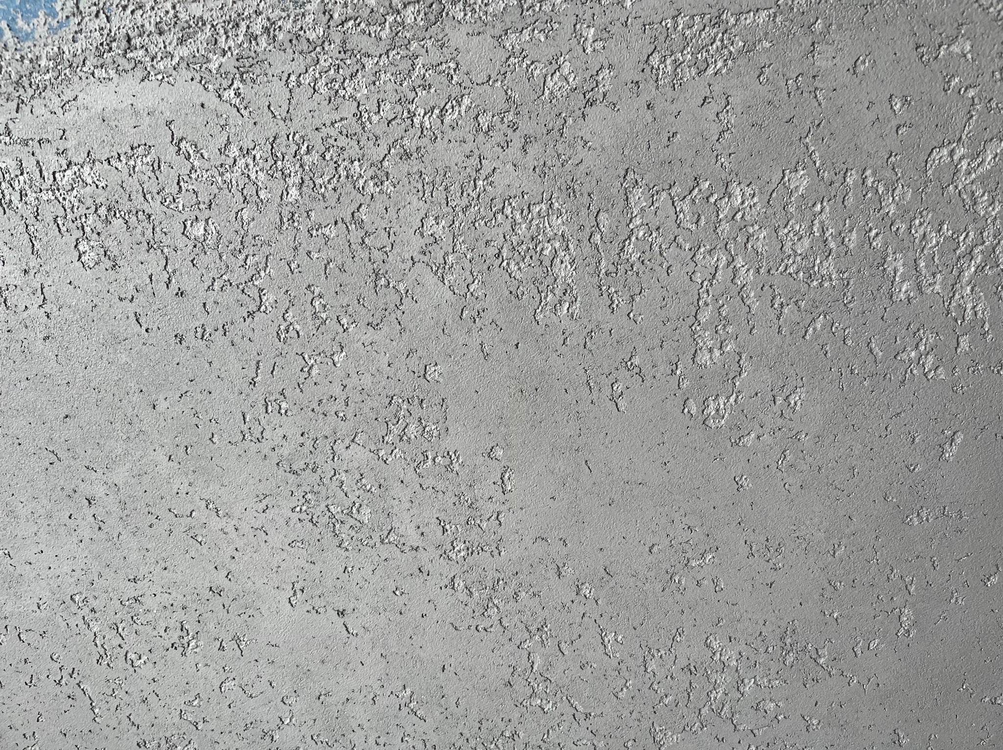 Szara ściana z wyraźną, nieregularną fakturą, przypominającą efekt dekoracyjnego tynku. Widoczne drobne nierówności i załamania światła podkreślające strukturę.