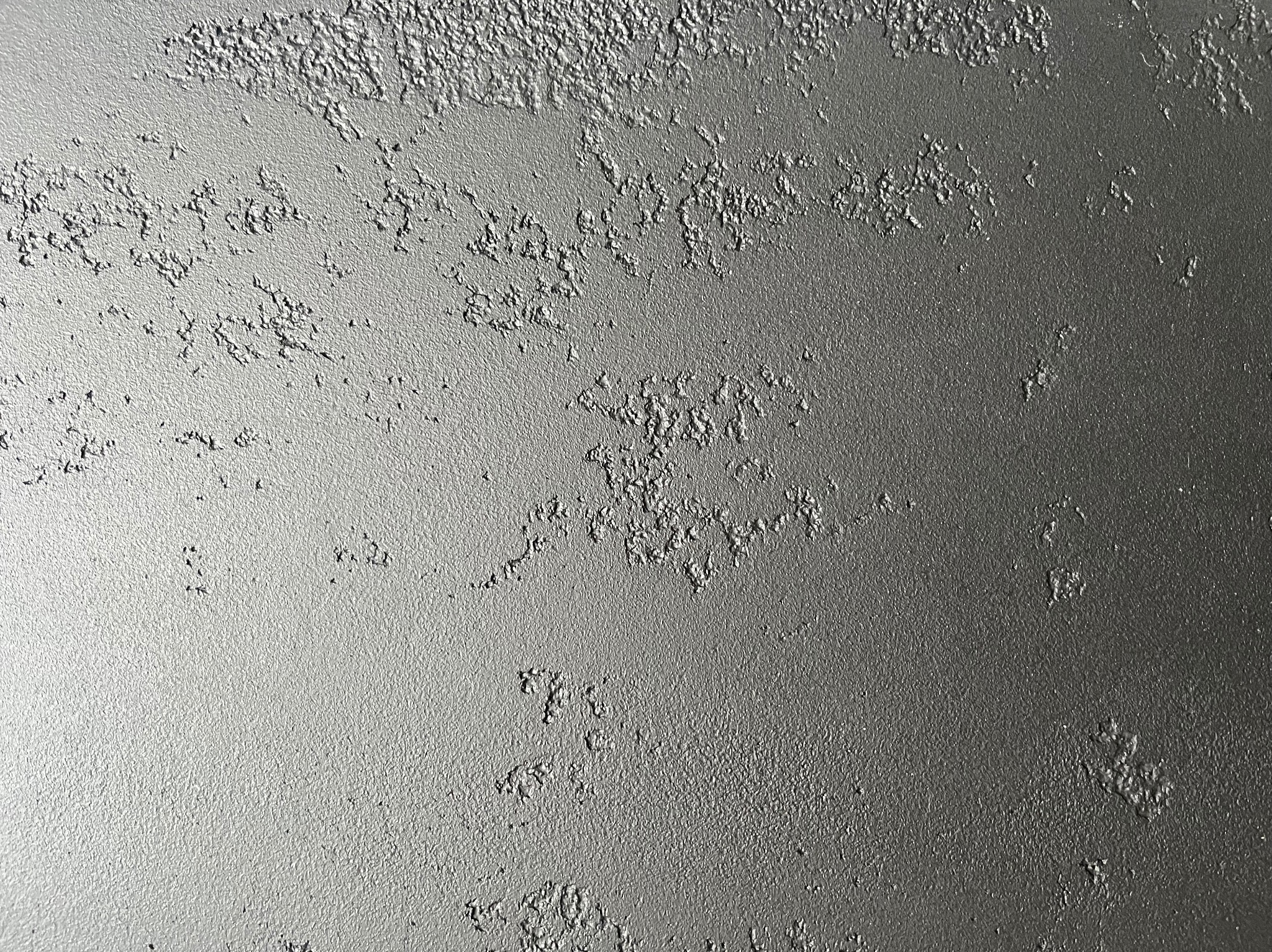 Szara ściana z wyraźną, nieregularną fakturą, przypominającą efekt dekoracyjnego tynku. Oświetlenie podkreśla nierówności powierzchni.