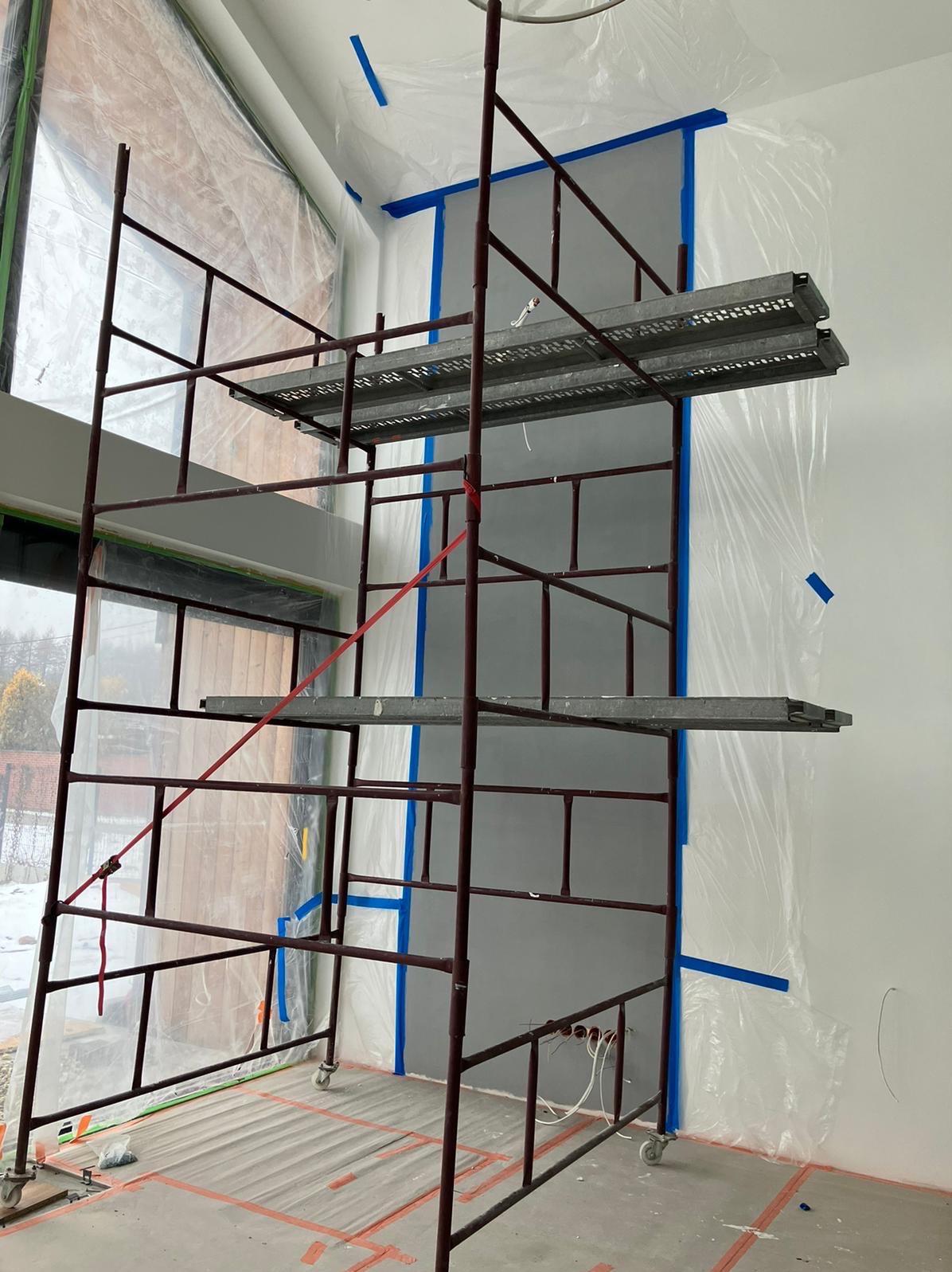 Wnętrze w trakcie malowania: rusztowanie na tle ściany zabezpieczonej folią i taśmą malarską. Widoczne okno i przygotowane podłoże. Etap prac wykończeniowych.