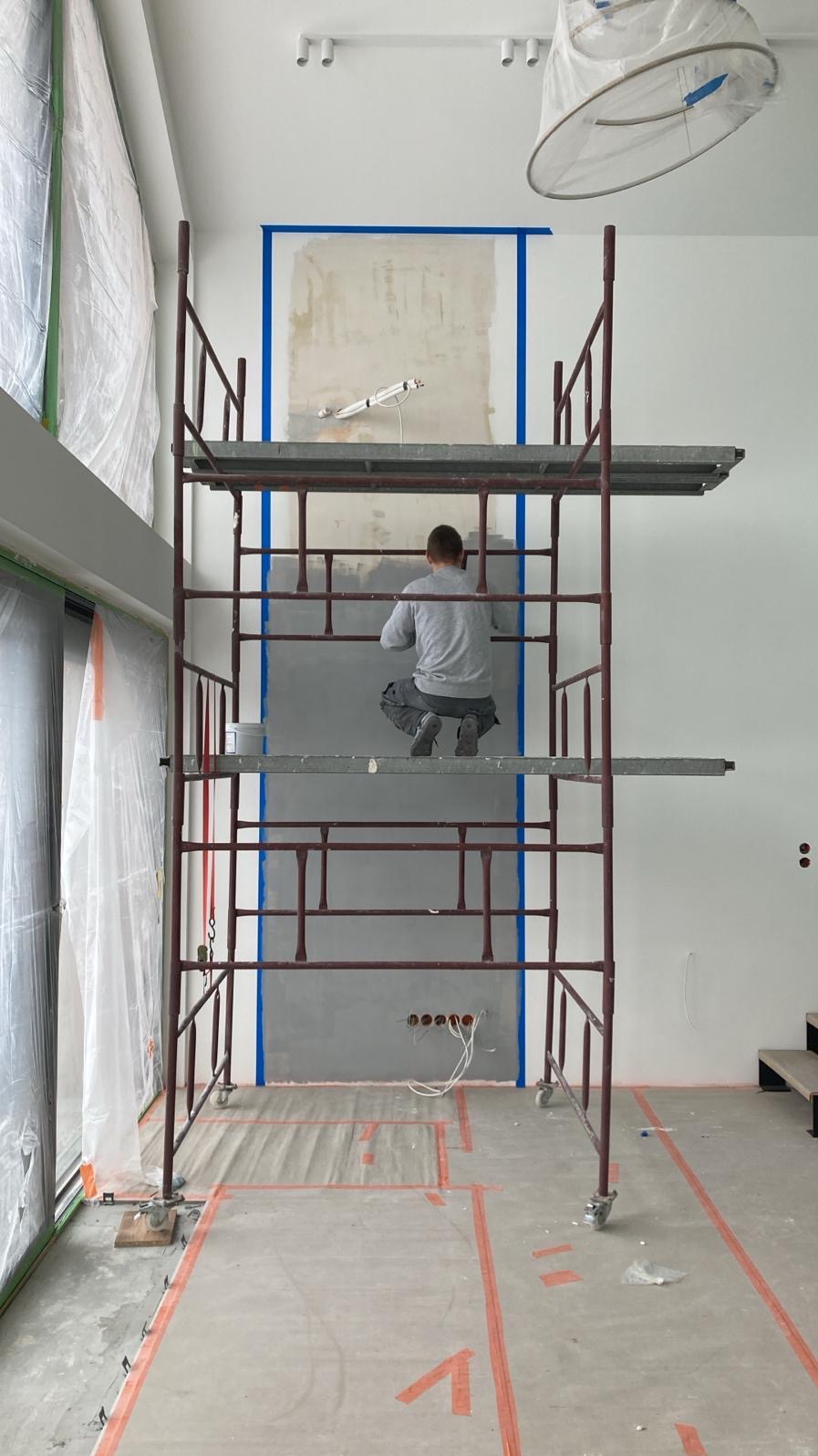Pracownik na rusztowaniu szpachluje ścianę w pomieszczeniu z dużymi oknami, zabezpieczonymi folią. Na podłodze widoczne taśmy malarskie.