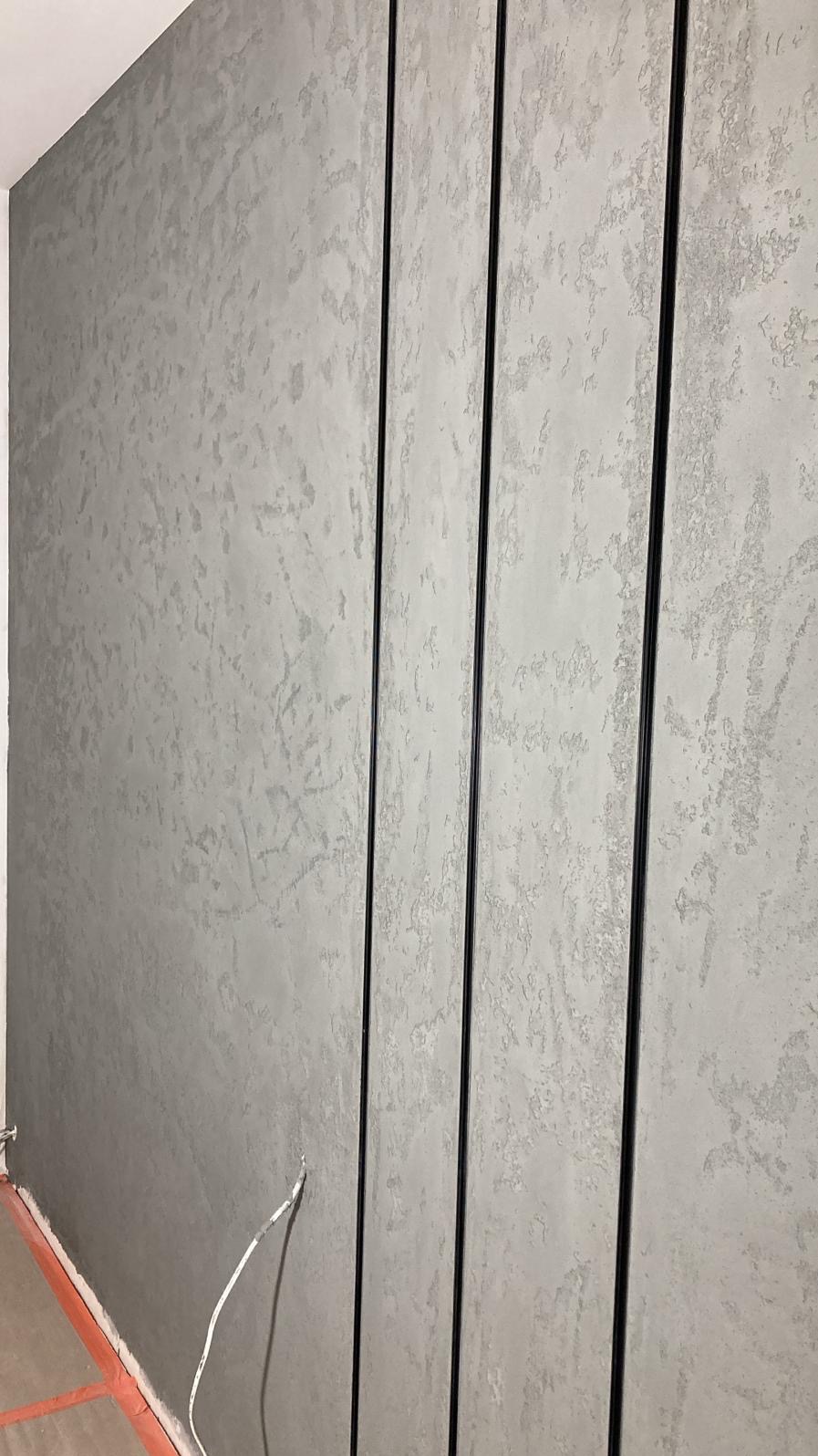 Szara ściana z dekoracyjnym tynkiem i czarnymi listwami pionowymi. Widoczny fragment podłogi z pomarańczową listwą przypodłogową i wystający kabel.