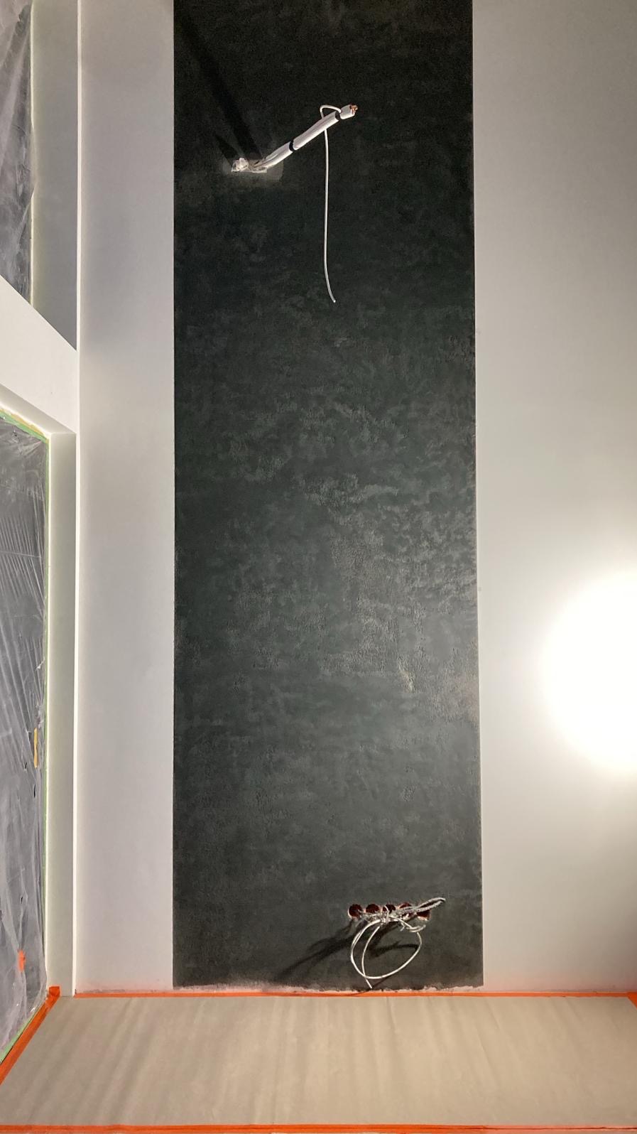 Wąski panel dekoracyjny na ścianie w odcieniach szarości, z widocznymi przewodami elektrycznymi u góry i dołu. Podłoga zabezpieczona folią malarską.