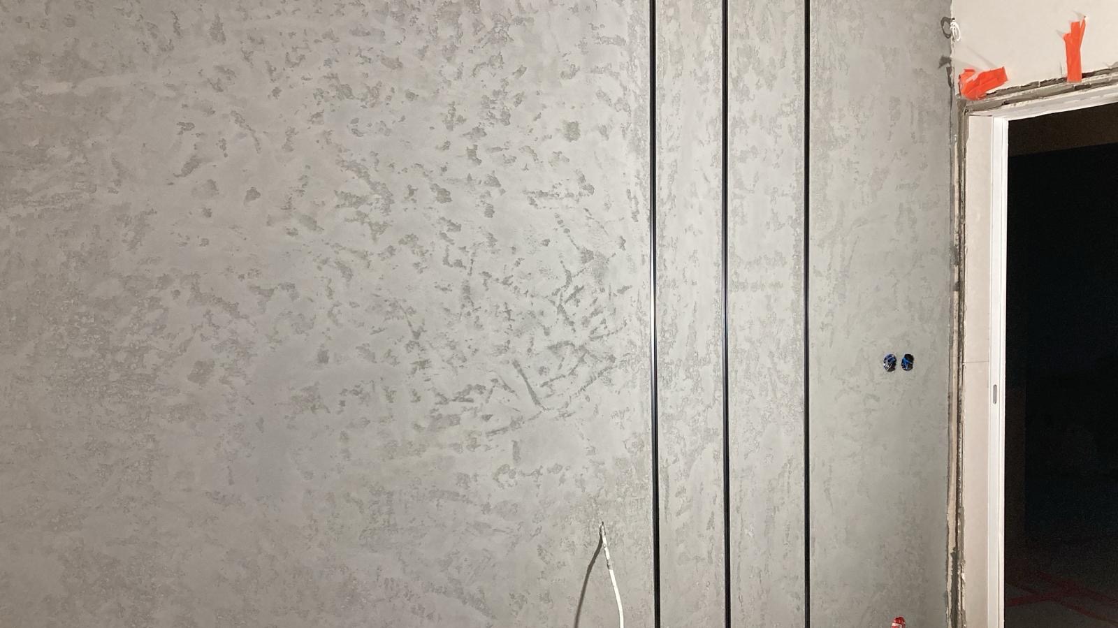 Ściana z dekoracyjnym, szarym tynkiem strukturalnym i pionowymi, czarnymi listwami. Widoczne gniazdka elektryczne i fragment ościeżnicy drzwi.