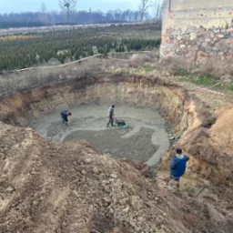 Rozległy wykop w ziemi z pracującymi robotnikami, ubijarka wibracyjna w użyciu, w tle szkółka drzew iglastych i fragment kamiennego muru.