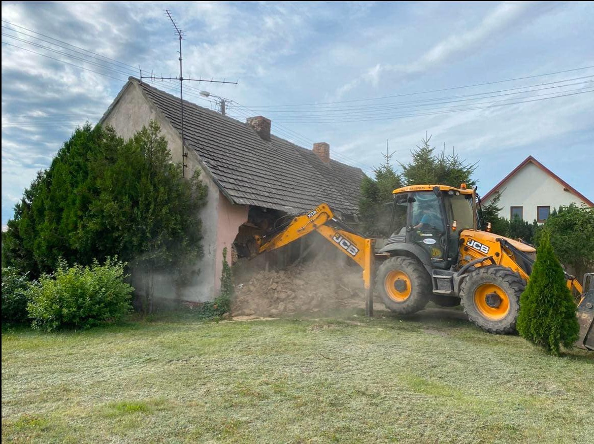 Żółta koparko-ładowarka JCB narusza ścianę starego, jednopiętrowego domu z ceglanym dachem, otoczonego zielenią, w tle widoczny kolejny budynek.