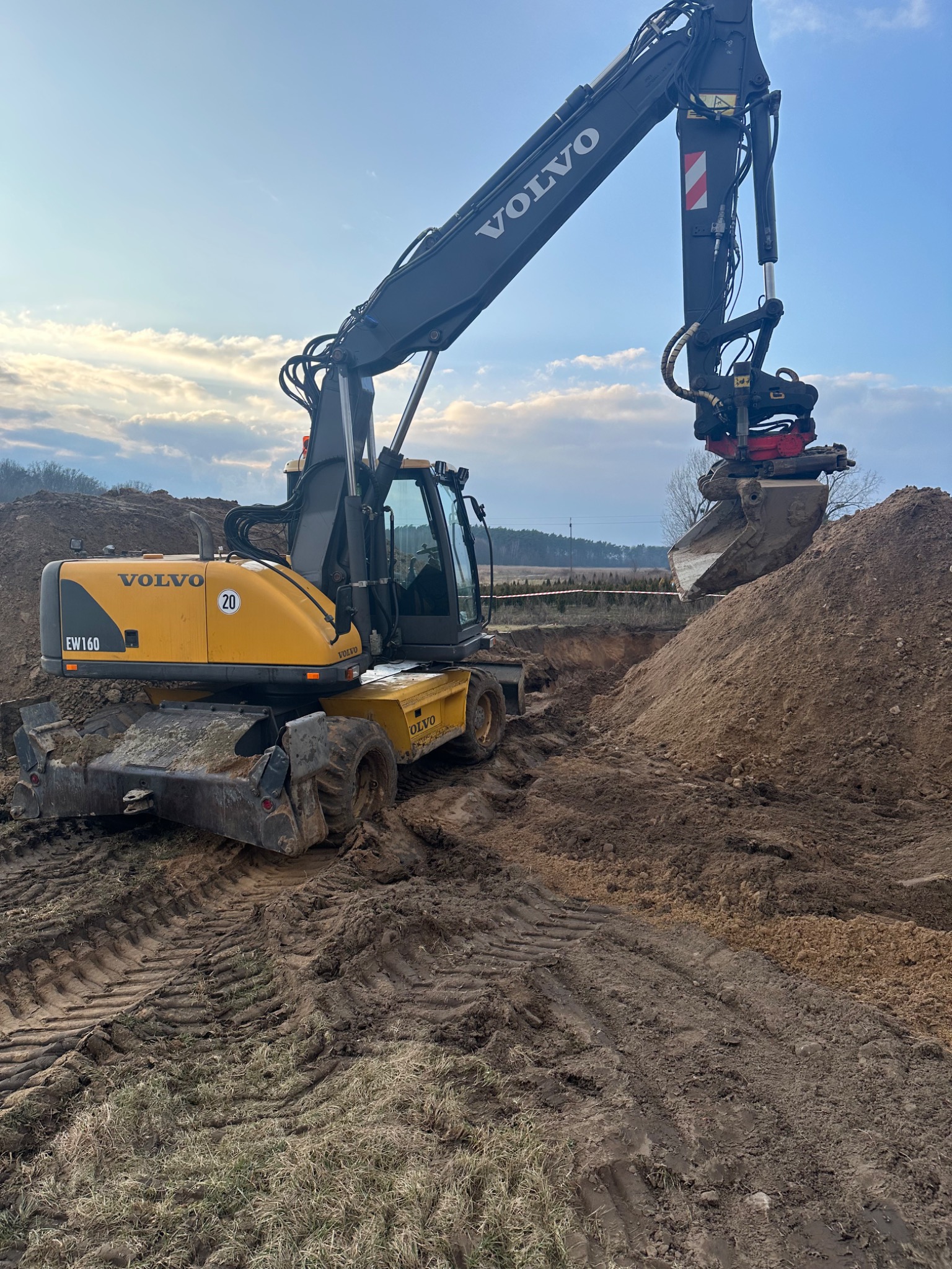 Żółta koparka kołowa Volvo EW160 z obrotową głowicą kopiąca ziemię, widoczne ślady opon na ziemi, w tle błękitne niebo z chmurami.
