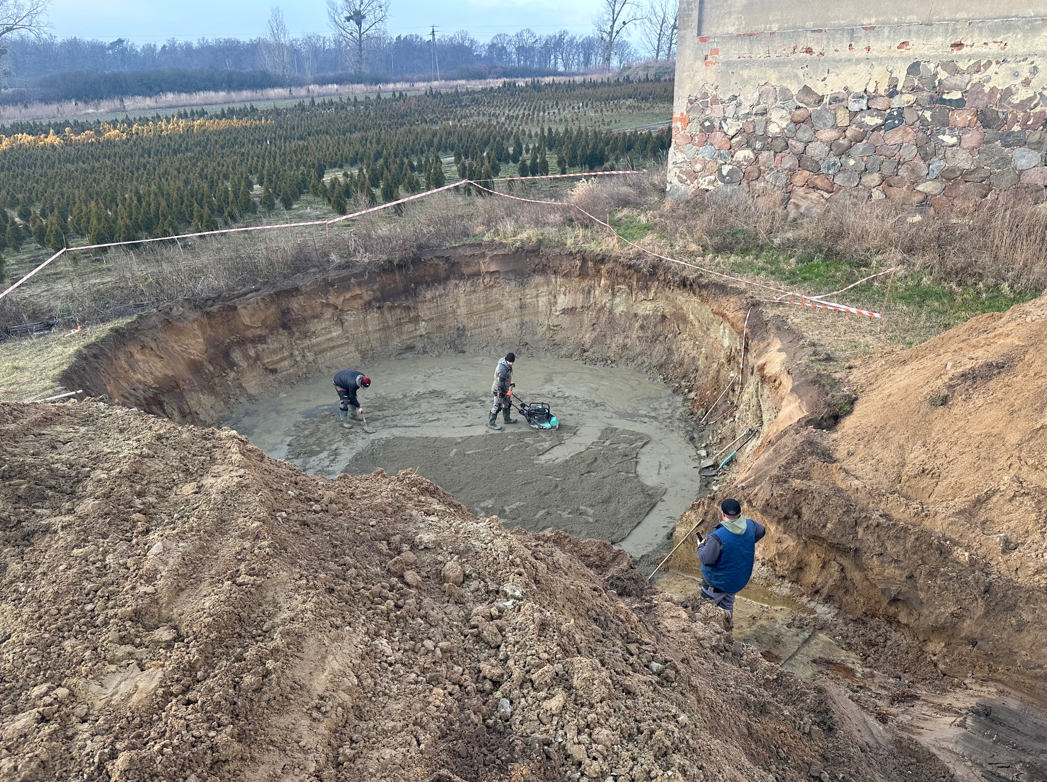 Rozległy wykop w ziemi z pracującymi robotnikami, ubijarka wibracyjna w użyciu, w tle szkółka drzew iglastych i fragment kamiennego muru.