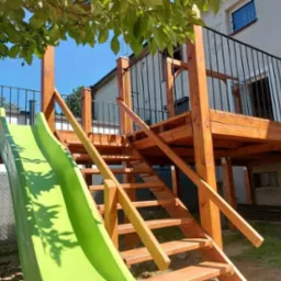 Drewniany taras z balustradą i schodami, zintegrowany z zieloną zjeżdżalnią dla dzieci, widok z dołu na tle budynku mieszkalnego.