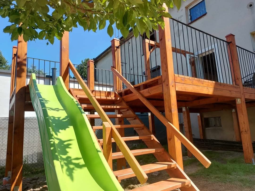 Drewniany taras z balustradą i schodami, zintegrowany z zieloną zjeżdżalnią dla dzieci, widok z dołu na tle budynku mieszkalnego.