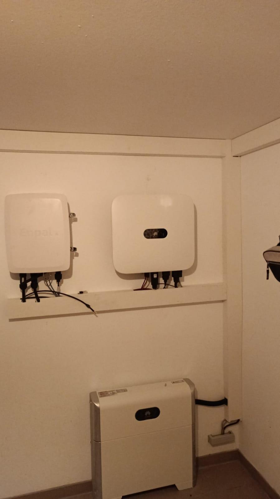 Dwa białe inwertery fotowoltaiczne zamontowane na białej ścianie wewnątrz pomieszczenia, z widocznym okablowaniem i dodatkowym urządzeniem na podłodze, również w białej obudowie.