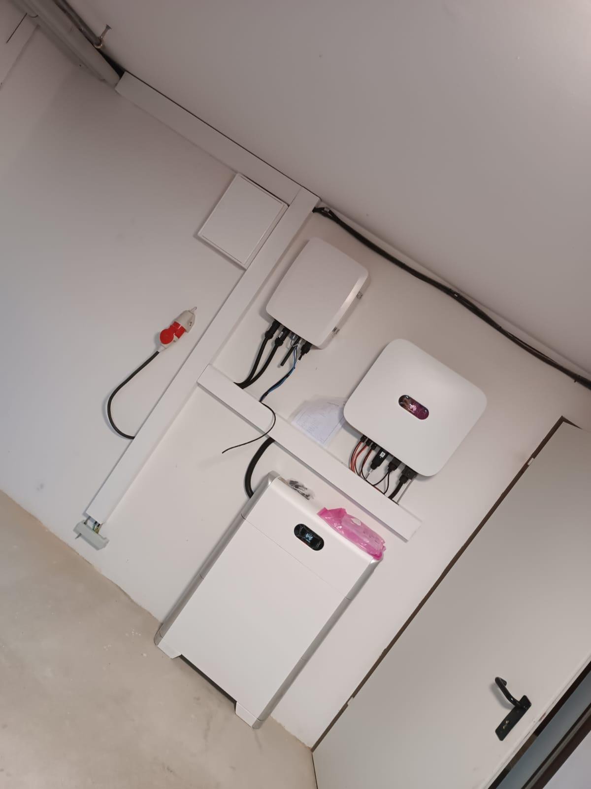 Wewnętrzna instalacja fotowoltaiczna: dwa białe inwertery i bateria podłączone do ściany za pomocą białych listew, widoczne kable i zabezpieczenia, różowa folia na baterii.