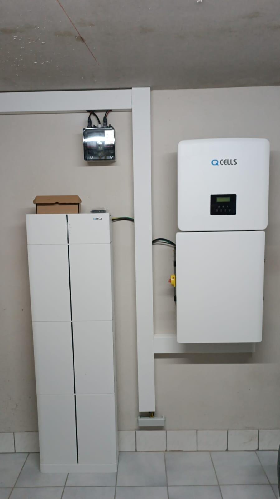 Biały akumulator energii Q.CELLS i inwerter zamontowane na ścianie w pomieszczeniu z wyeksponowanymi przewodami i elementami instalacji elektrycznej.